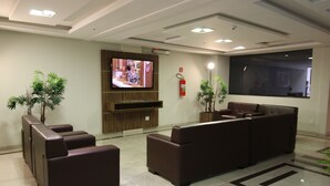 A lobby with a large flat-screen TV, brown leather seating, potted plants, and a fire extinguisher on the wall.