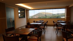 A dining area with multiple tables and chairs, large windows with a view of hills and a river, and a wooden cabinet on the left.