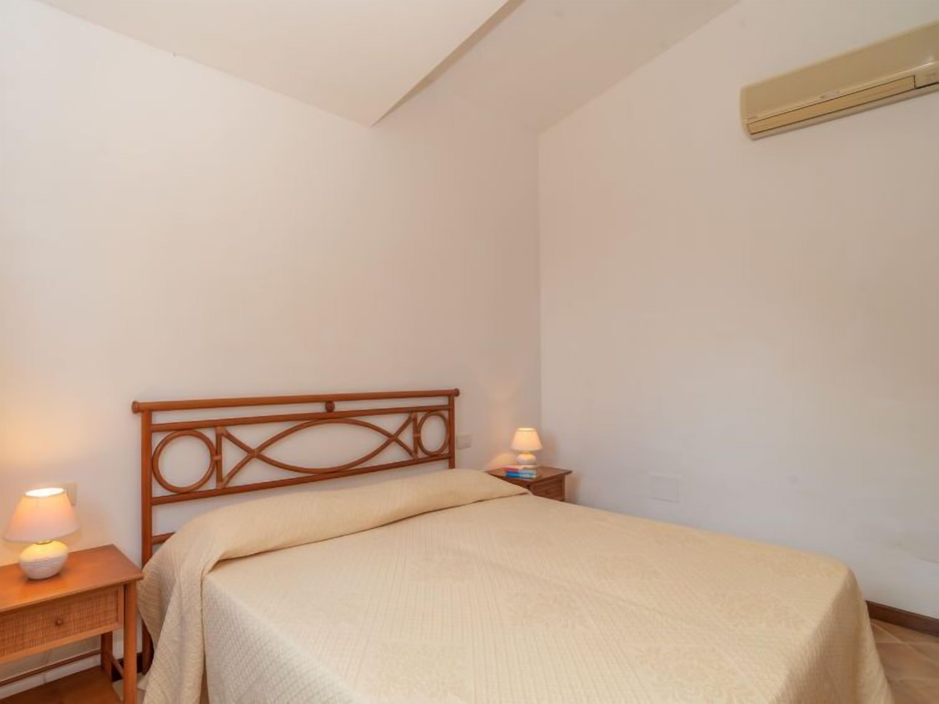 A bedroom with a wooden headboard, a bed with a beige blanket, two bedside tables with lamps, and a wall-mounted air conditioning unit.