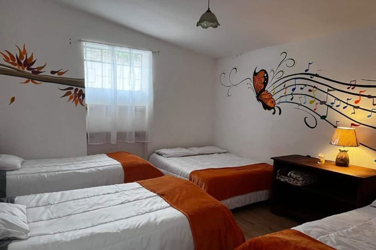 A hotel room with three beds, a window with sheer curtains, a wall mural of a butterfly, and a bedside table with a lamp.