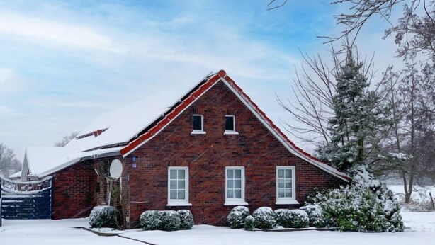 A brick house with a steep roof, surrounded by snow-covered ground and trees.