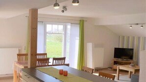 A modern kitchen with a central island, wooden chairs, a dining table, a TV, and a window with curtains.