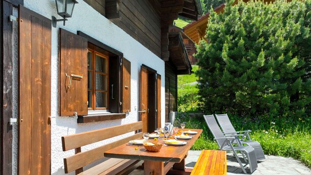An outdoor dining area with wooden benches and tables, surrounded by greenery and a building with wooden shutters. - Notre Rêve by Interhome (Zinal)