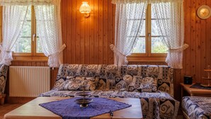 A cozy cabin interior with wooden walls, a patterned sofa, a table with a decorative centerpiece, and two windows with curtains. - Notre Rêve by Interhome (Zinal)
