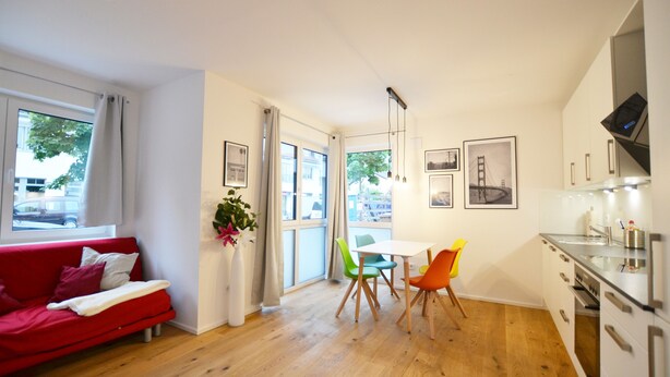A modern living room with a red sofa, a dining area with a white table and colorful chairs, and a kitchen with white cabinets and a stainless steel range hood. - Modern apartment near downtown (Munich)