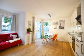 A modern living room with a red sofa, a dining area with a white table and colorful chairs, and a kitchen with white cabinets and a stainless steel range hood. - Modern apartment near downtown (Munich)
