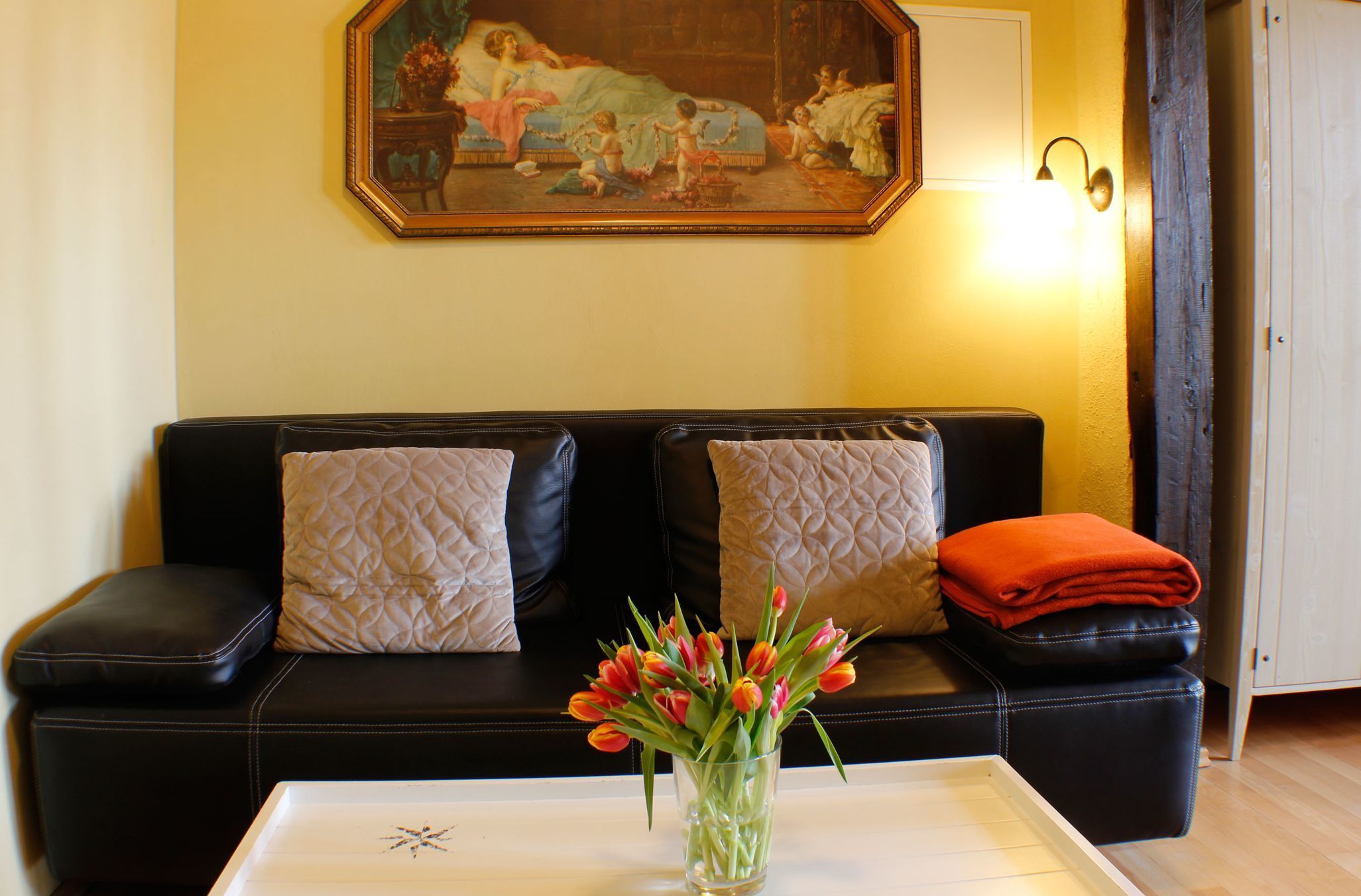 Ein Wohnzimmer mit einem schwarzen Ledersofa, zwei gemusterten Kissen, einer Glasvase mit roten Tulpen und einem Gemälde an der Wand.