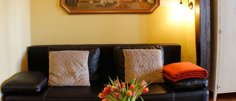 Ein Wohnzimmer mit einem schwarzen Ledersofa, zwei gemusterten Kissen, einer Glasvase mit roten Tulpen und einem Gemälde an der Wand.