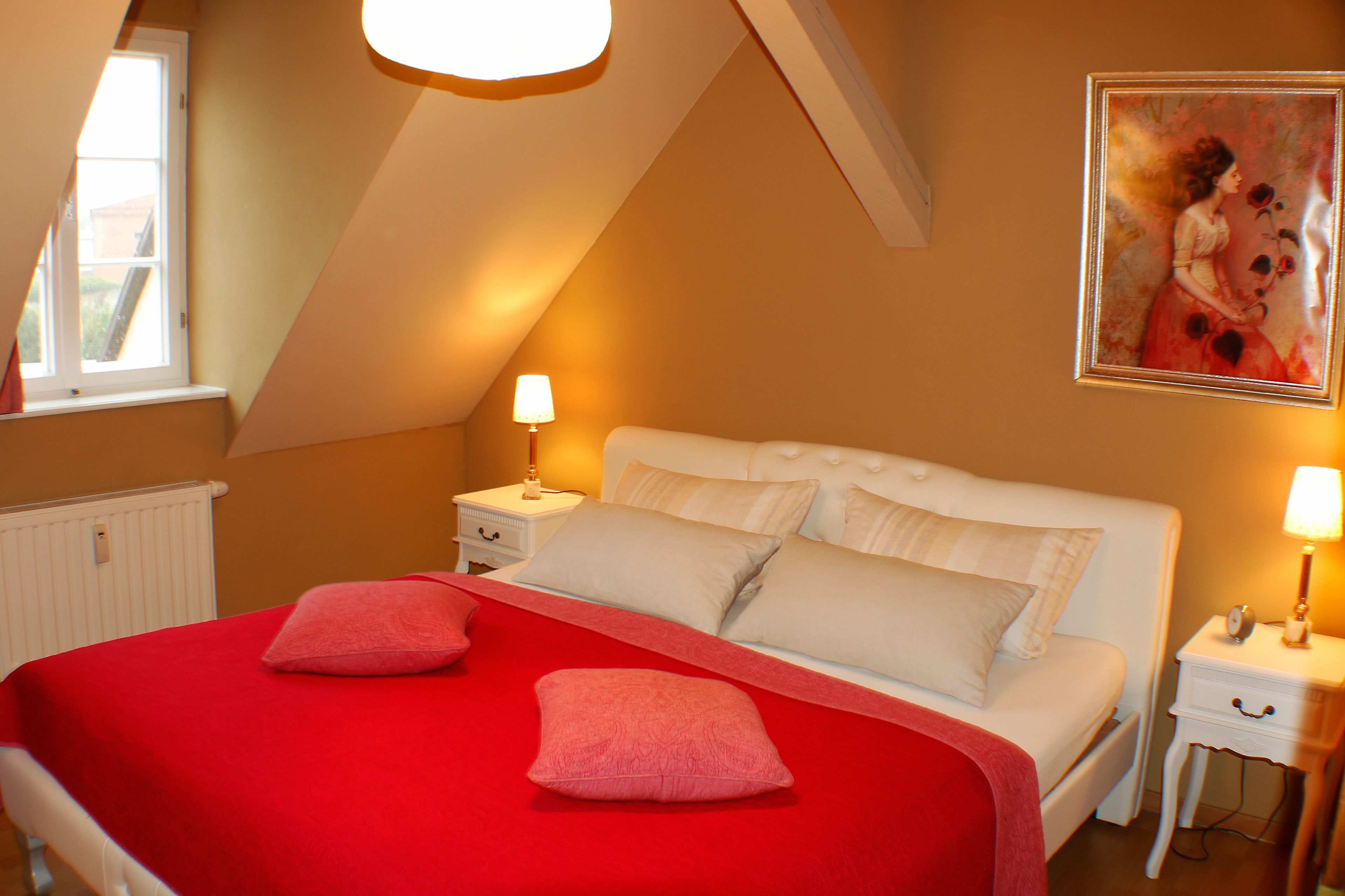 A bedroom with a sloped ceiling, a bed with a red bedspread and two pink pillows, white nightstands with lamps, and a framed painting on the wall.