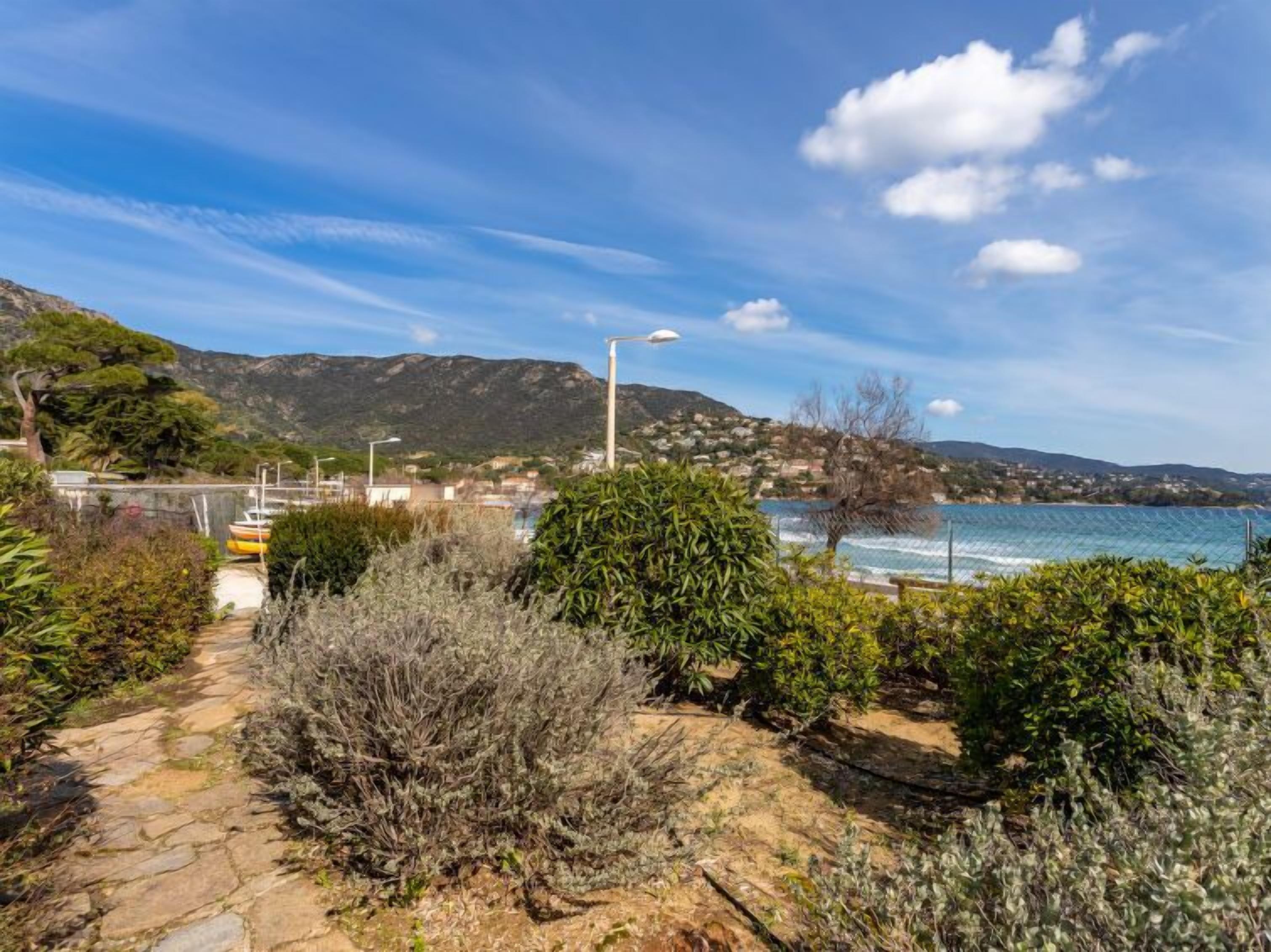 A coastal path with a stone walkway, dense shrubbery, and a view of the sea and distant hills.