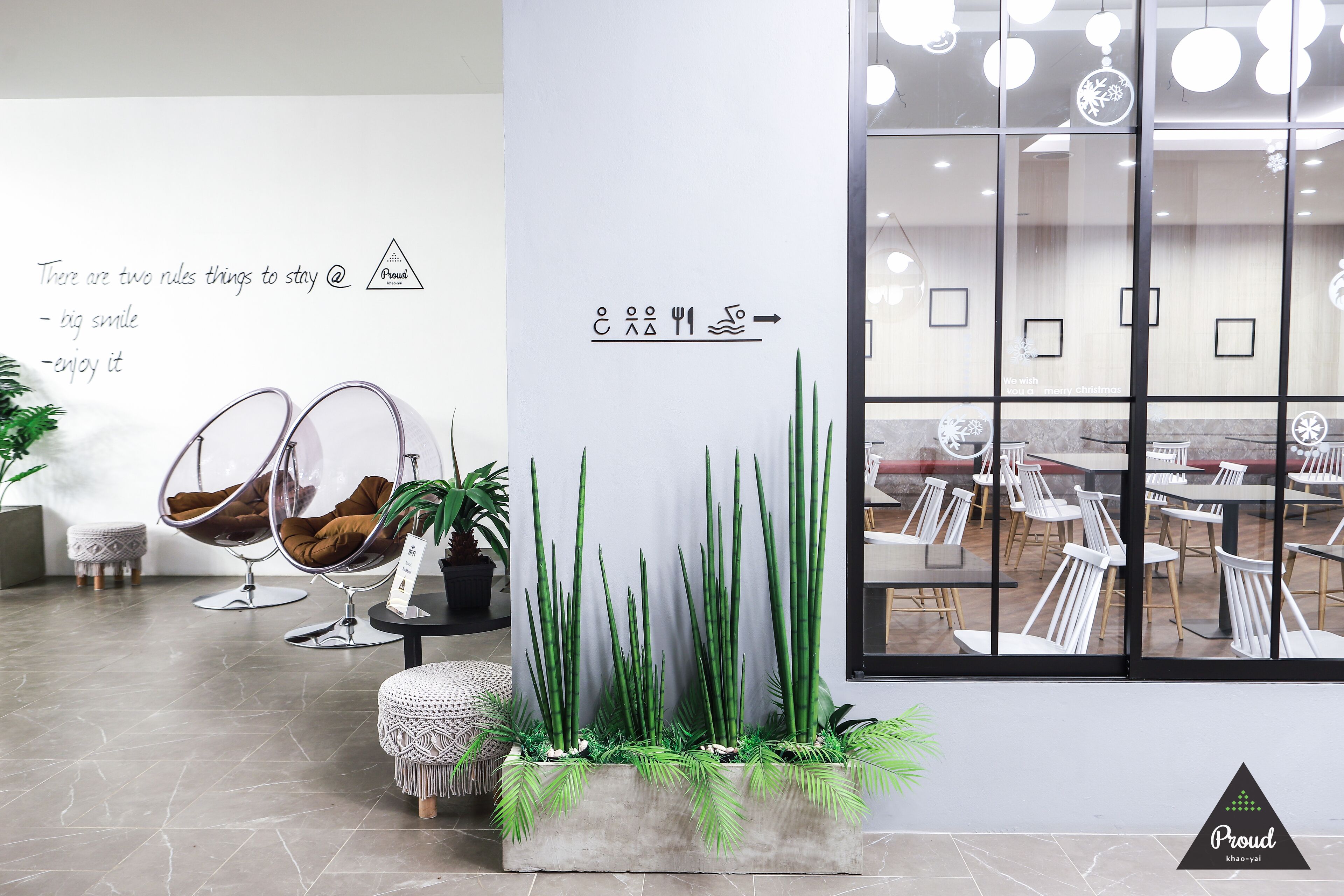 A modern interior with a seating area featuring two egg chairs, a small round table, and a potted plant. There is a wall with a sign and a large window showing a dining area.