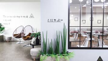 Un interior moderno con una zona de estar que incluye dos sillones huevo, una mesita redonda y una planta en maceta. Se ve una pared con un cartel y un ventanal grande que muestra un comedor.