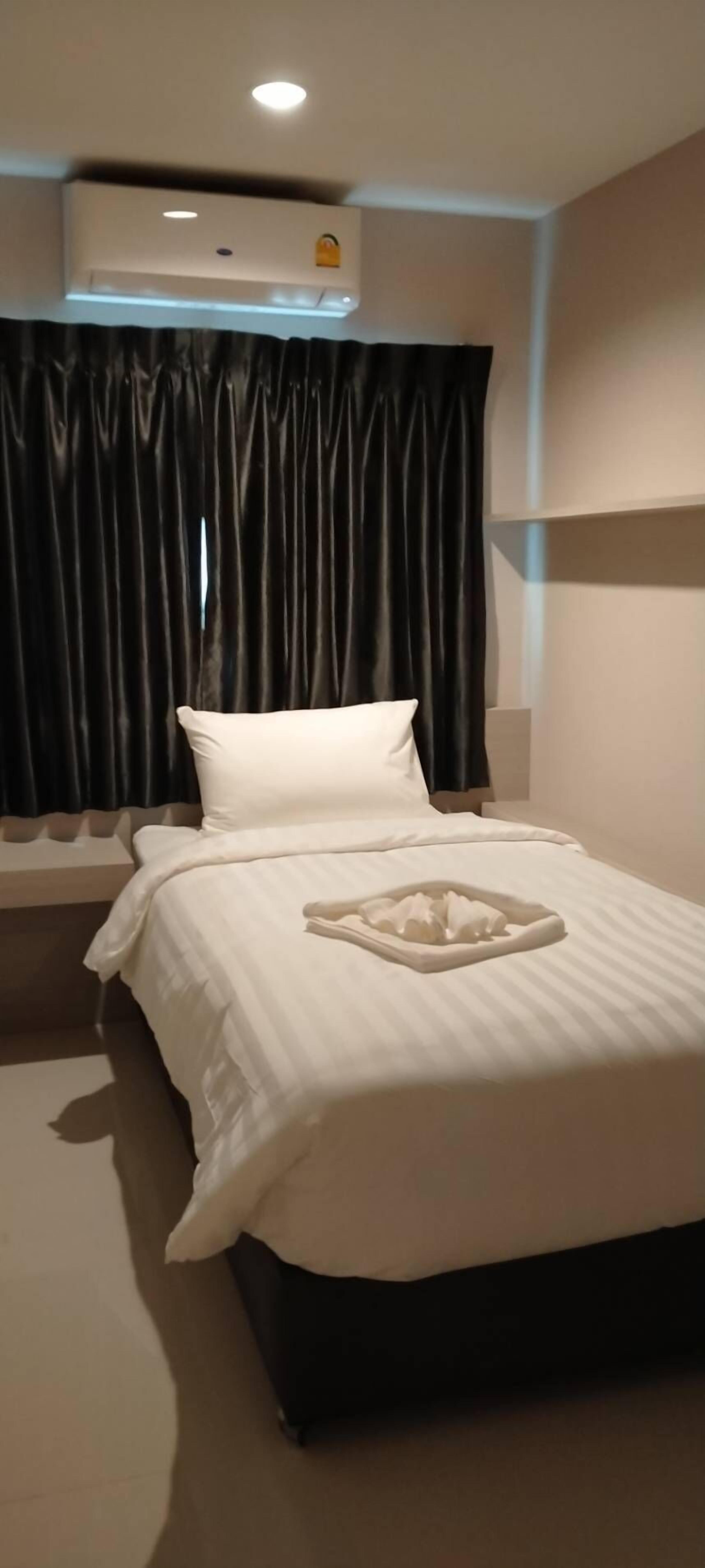 A hotel room with a bed, a window with dark curtains, an air conditioning unit, and a bedside table.