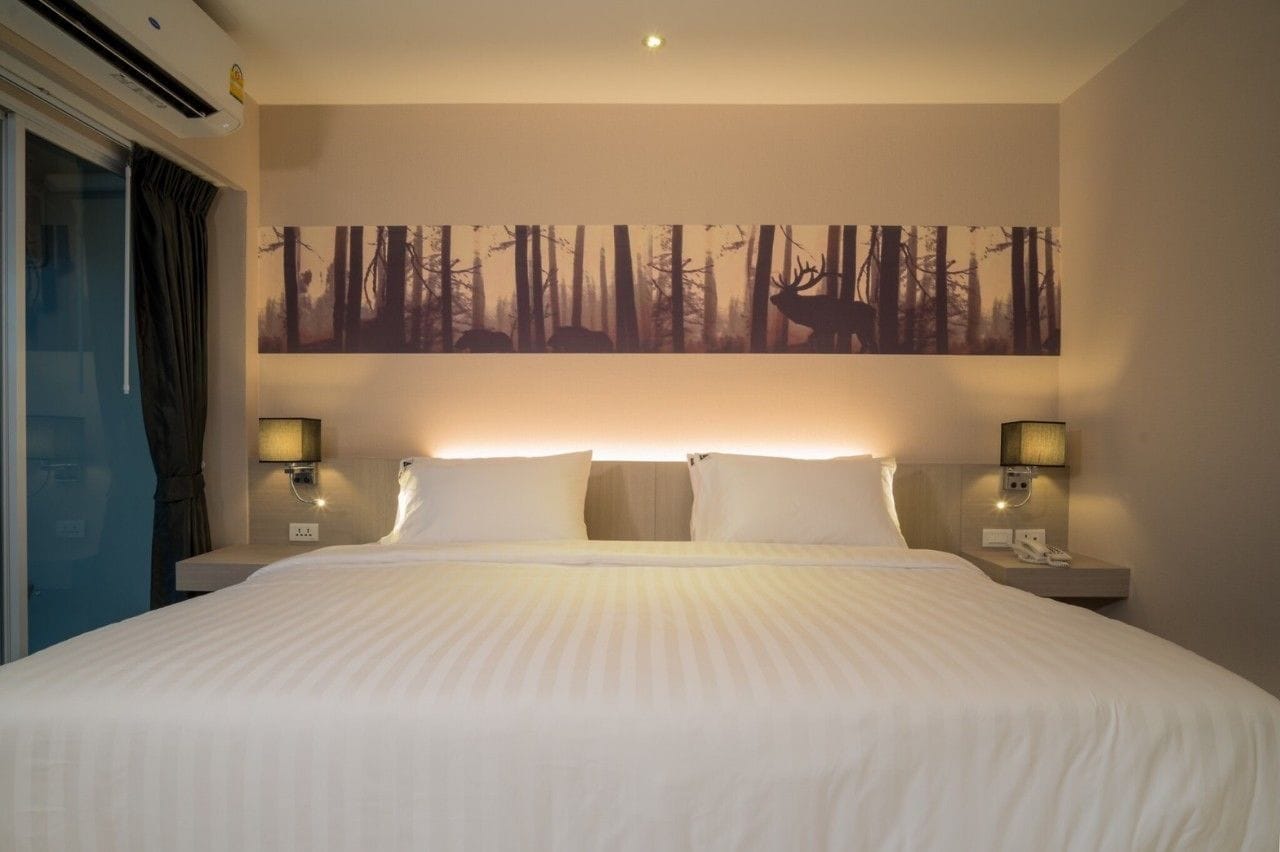 Hotelkamer met een groot bed, twee bedlampjes, een wanddecoratie van een bos en een raam met gordijnen.