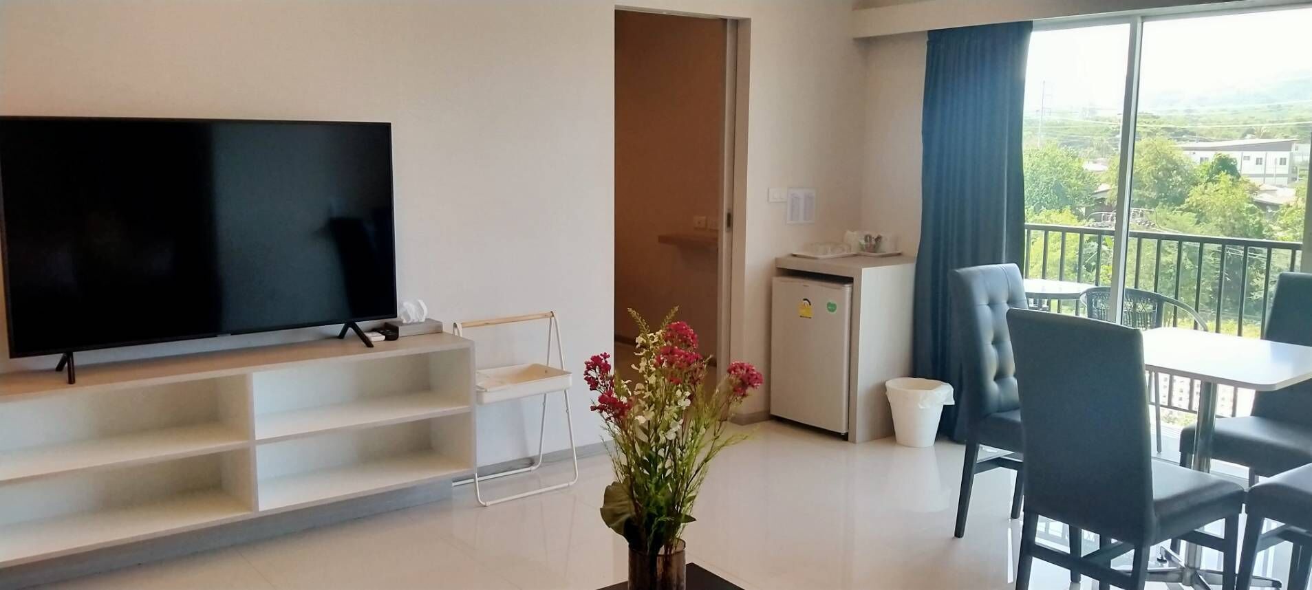 A modern hotel room with a large flat-screen TV, a dining area with a table and chairs, a small kitchenette, and a balcony with a view of the surrounding landscape.