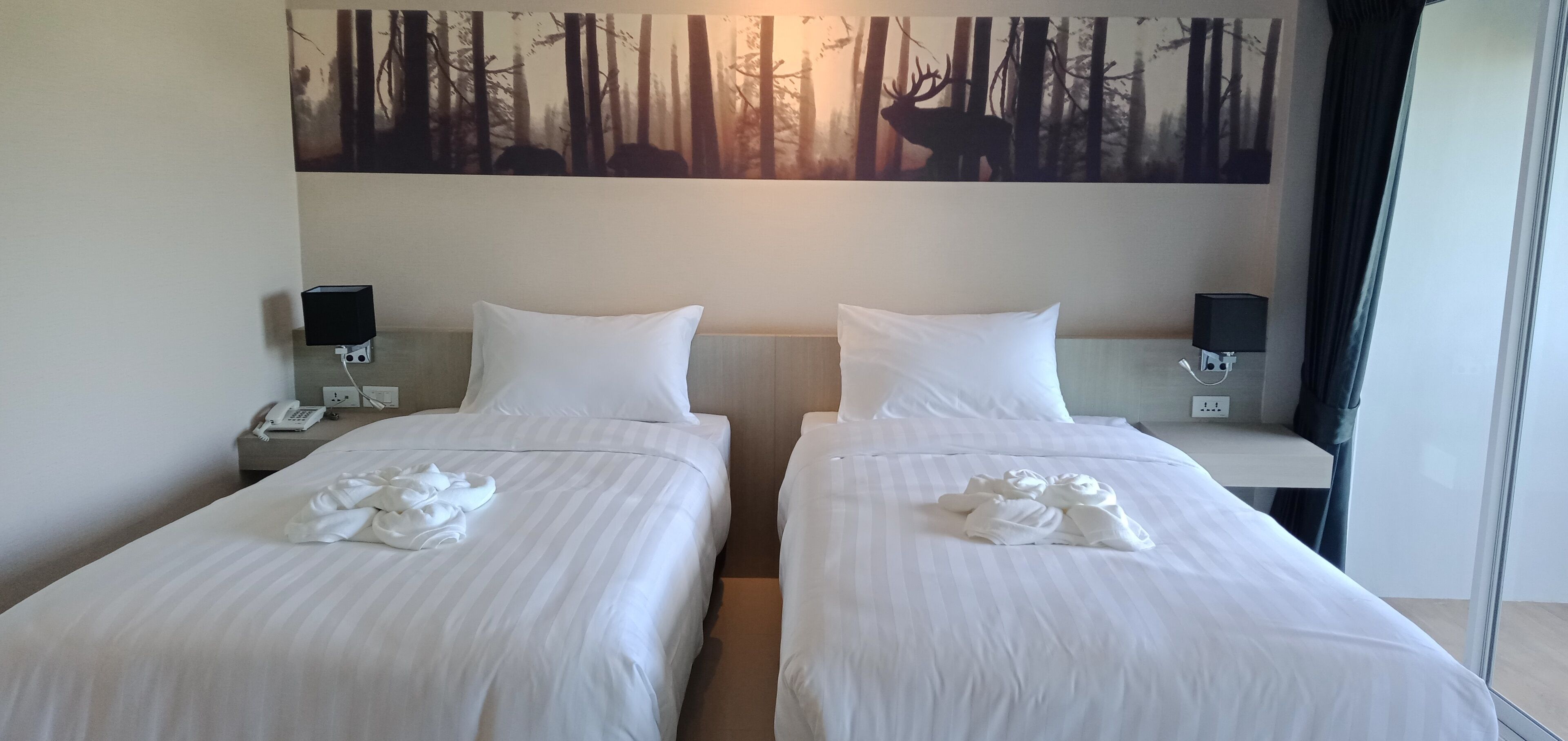 A hotel room with two beds, each with white linens and a forest-themed painting above. There are bedside tables with lamps and a small table with a phone and a speaker.