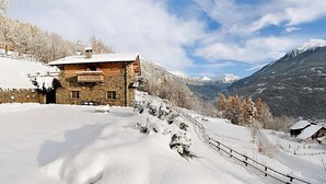 Un chalet in pietra con balcone in legno, circondato da montagne e alberi innevati.