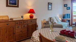 A cozy living room with a wooden cabinet, a red lamp, a sofa, a TV, and a dining table with a patterned tablecloth. - Victoria 1 C1 by Interhome (Nendaz)