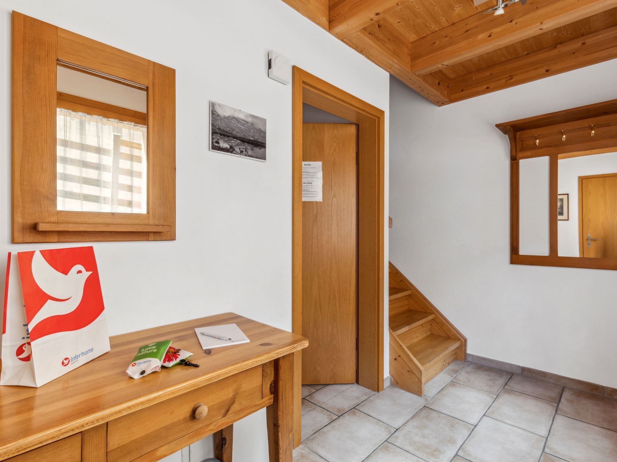 Une chambre d’hôtel avec un bureau en bois, une fenêtre avec des stores, un tableau encadré au mur, une porte donnant sur une autre pièce et un escalier en bois.
