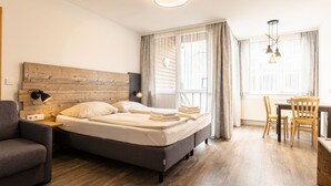 Ein modernes Schlafzimmer mit einem Holzkopfteil, einem grauen Sofa, einem Esstisch mit Stühlen und einem großen Fenster mit Vorhängen.
