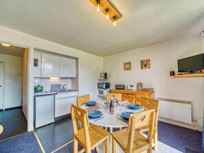 Une cuisine compacte avec des meubles blancs, un four à micro-ondes et un petit réfrigérateur. Une table à manger avec des assiettes bleues et des chaises en bois est dressée pour quatre personnes.