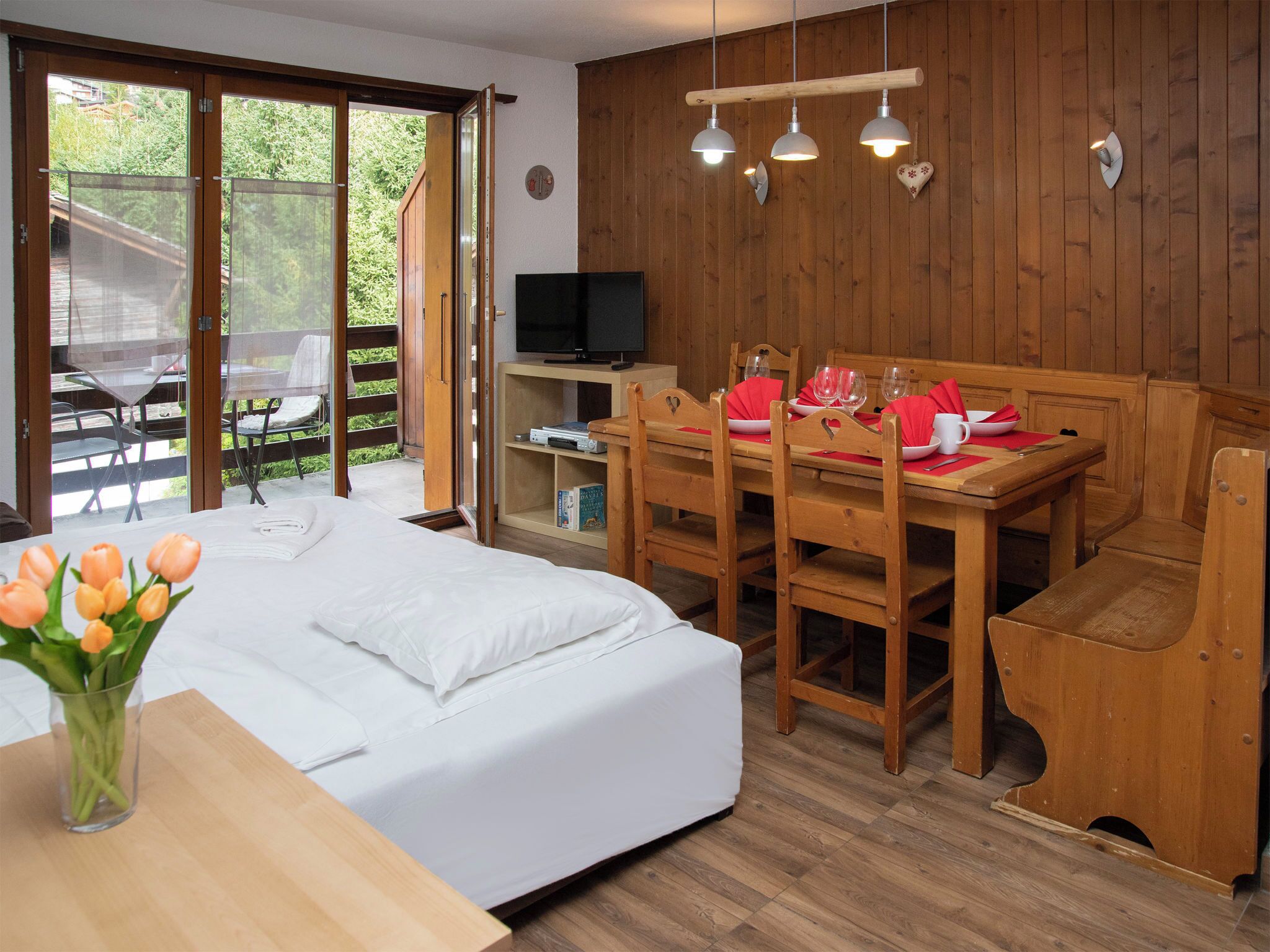 A bedroom with a bed, a dining table with chairs, a TV, and a wooden wall.