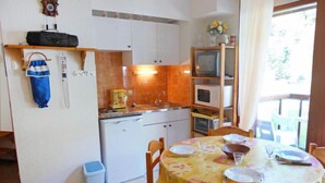 Een compacte keuken met witte kastjes, een magnetron en een koffiezetapparaat. Er is een eettafel met stoelen en een kleine prullenbak.
