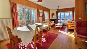 A cozy room with a dining table, chairs, a sofa, and a red couch. There are large windows with curtains, a fireplace, and a view of the mountains.