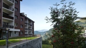 A multi-story building with balconies, a stone wall, and a grassy area with a tree.