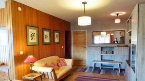 A cozy living room with a wooden wall, a sofa, a small table with a lamp, a bench, and a colorful striped rug.