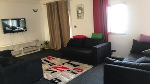 Un salon avec un téléviseur à écran plat fixé au mur, un canapé noir agrémenté de coussins rouges et verts, un tapis à motifs et une fenêtre avec des rideaux rouges.