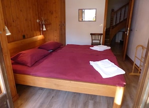 Uma cama de madeira com colcha vermelha, uma cadeira pequena e um espelho na parede.