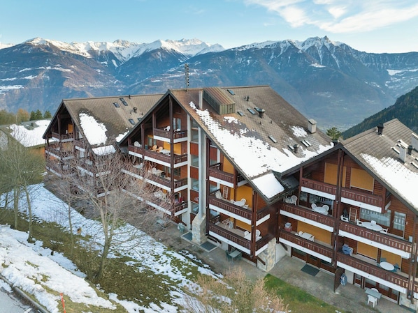 A multi-story wooden chalet with balconies, set against a backdrop of snow-covered mountains.