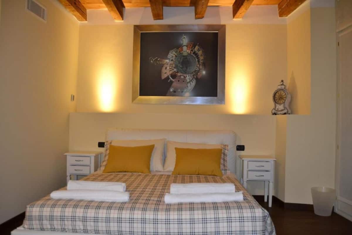 Quarto com cama, duas mesas de cabeceira, relógio na parede e uma obra de arte emoldurada sobre a cama.