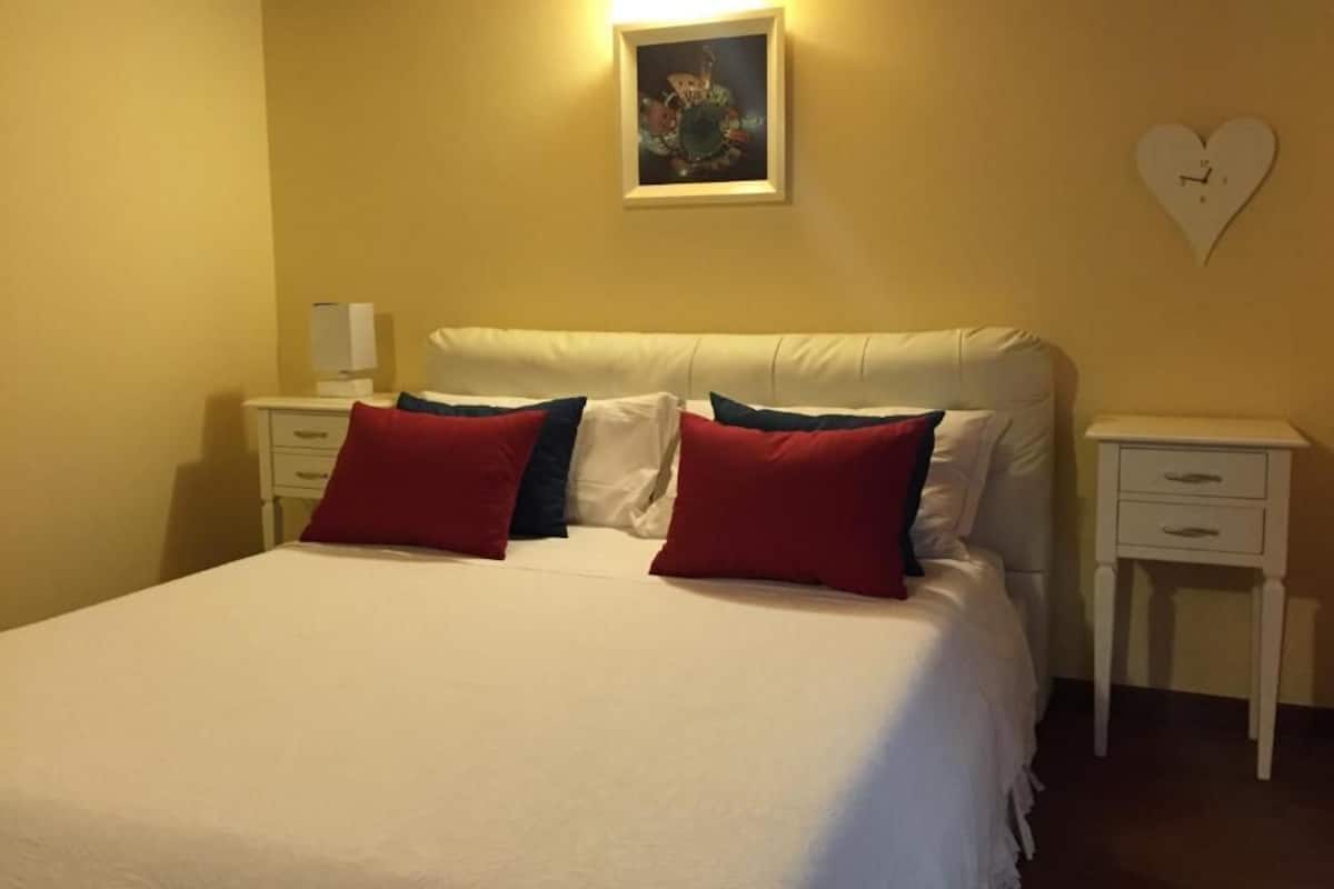 Cama bem arrumada com almofadas brancas e vermelhas, mesa de cabeceira com abajur e relógio de parede.