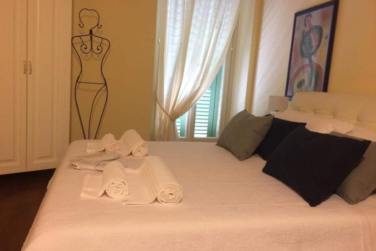 Cama bem arrumada com toalhas enroladas, cabeceira branca e um quadro na parede.