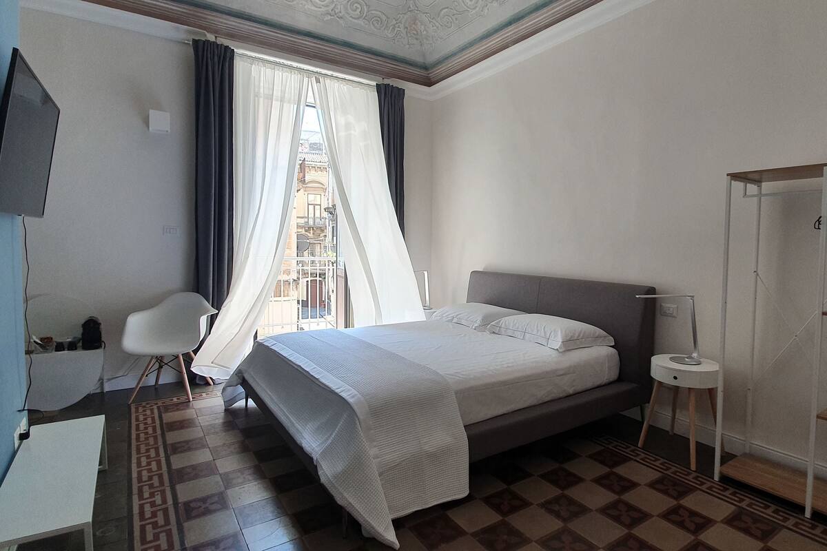 A hotel room with a bed, a desk, a chair, a TV, a wardrobe, and a window with curtains.
