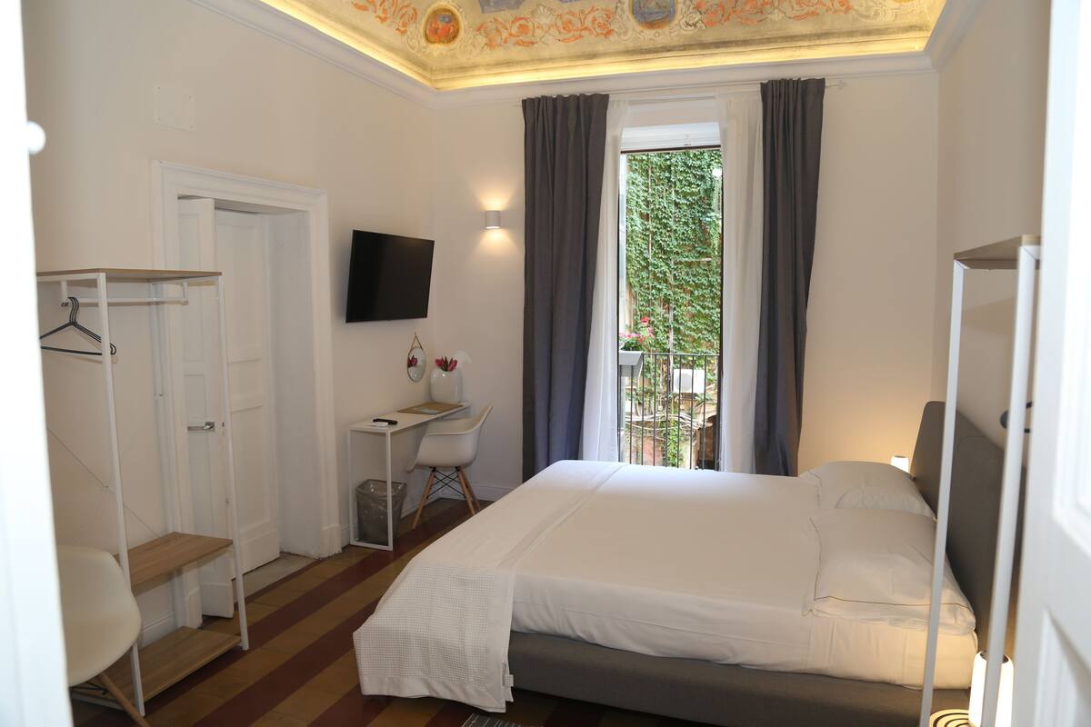A bedroom with a bed, a desk, a chair, a TV, and a window with curtains.