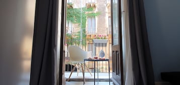 A view through a window with curtains, revealing a balcony with a white chair, a small table, and a potted plant.