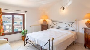 A bedroom with a bed, two bedside lamps, a radiator, a window with curtains, and a view of buildings outside.