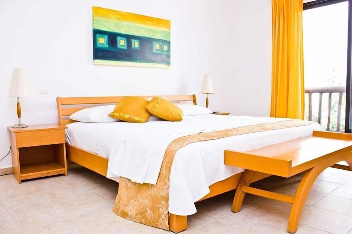 Quarto com cama de madeira, roupa de cama branca, duas luminárias de cabeceira, um banco ao pé da cama e um quadro na parede.