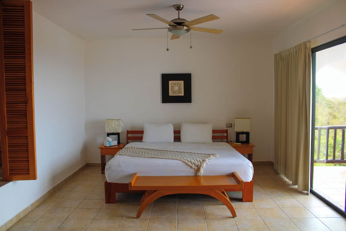 Um quarto com cama, mesas de cabeceira, ventoinha no teto, um quadro na parede e vista para o exterior pela janela.