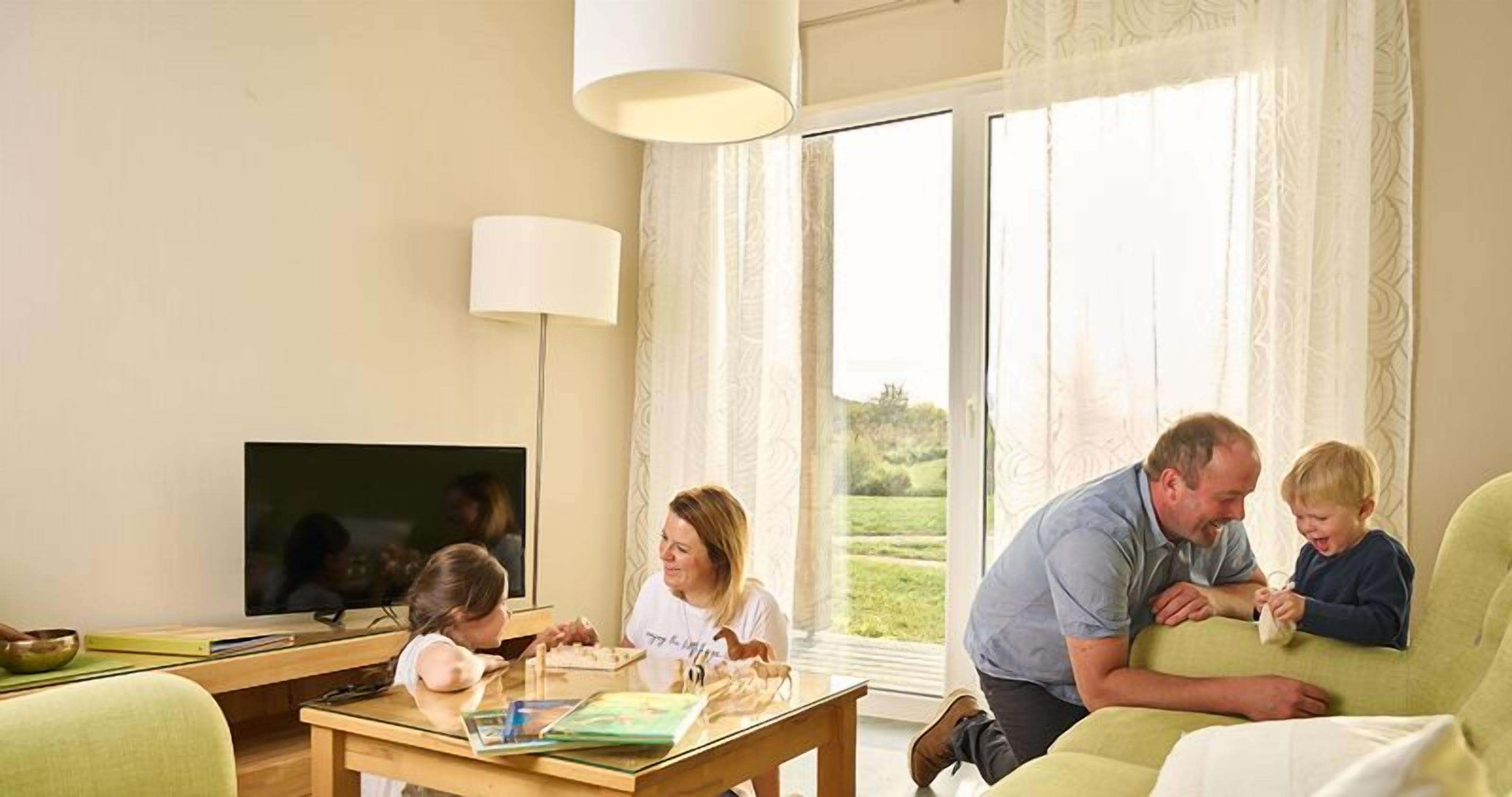 A family in a living room with a TV, a table with books and a bowl, and a large window with curtains.
