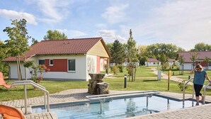 Una piscina con una escalera metálica, rodeada de una zona pavimentada y un pequeño jardín. Hay sillones de descanso y un edificio con techo rojo al fondo.