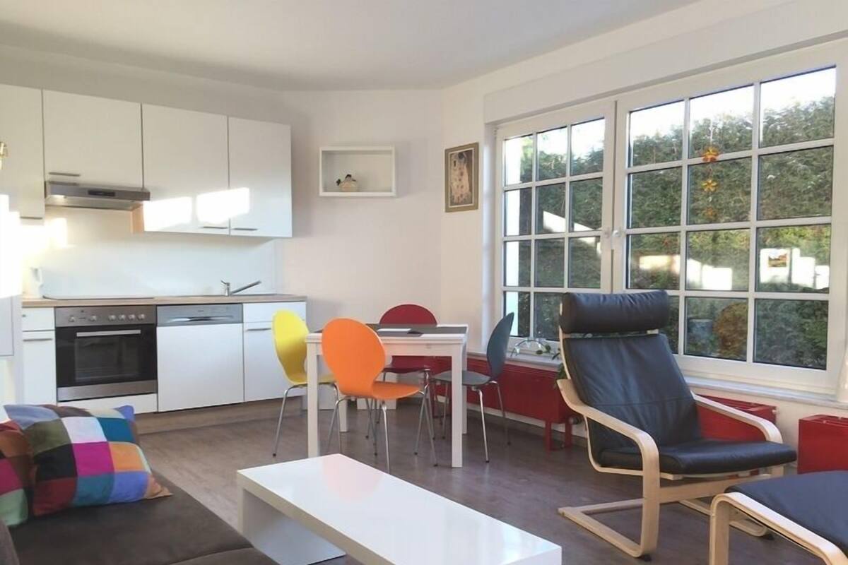Eine moderne Küche mit weißen Schränken, einem eingebauten Ofen und einem Geschirrspüler. Ein Essbereich mit weißem Tisch, bunten Stühlen und einem schwarzen Ledersessel. Ein großes Fenster bietet Blick auf Bäume draußen.