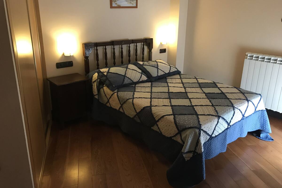 Un dormitorio con piso de madera, una cama con manta a cuadros, dos lámparas de noche, una mesita de noche y un cuadro en la pared.