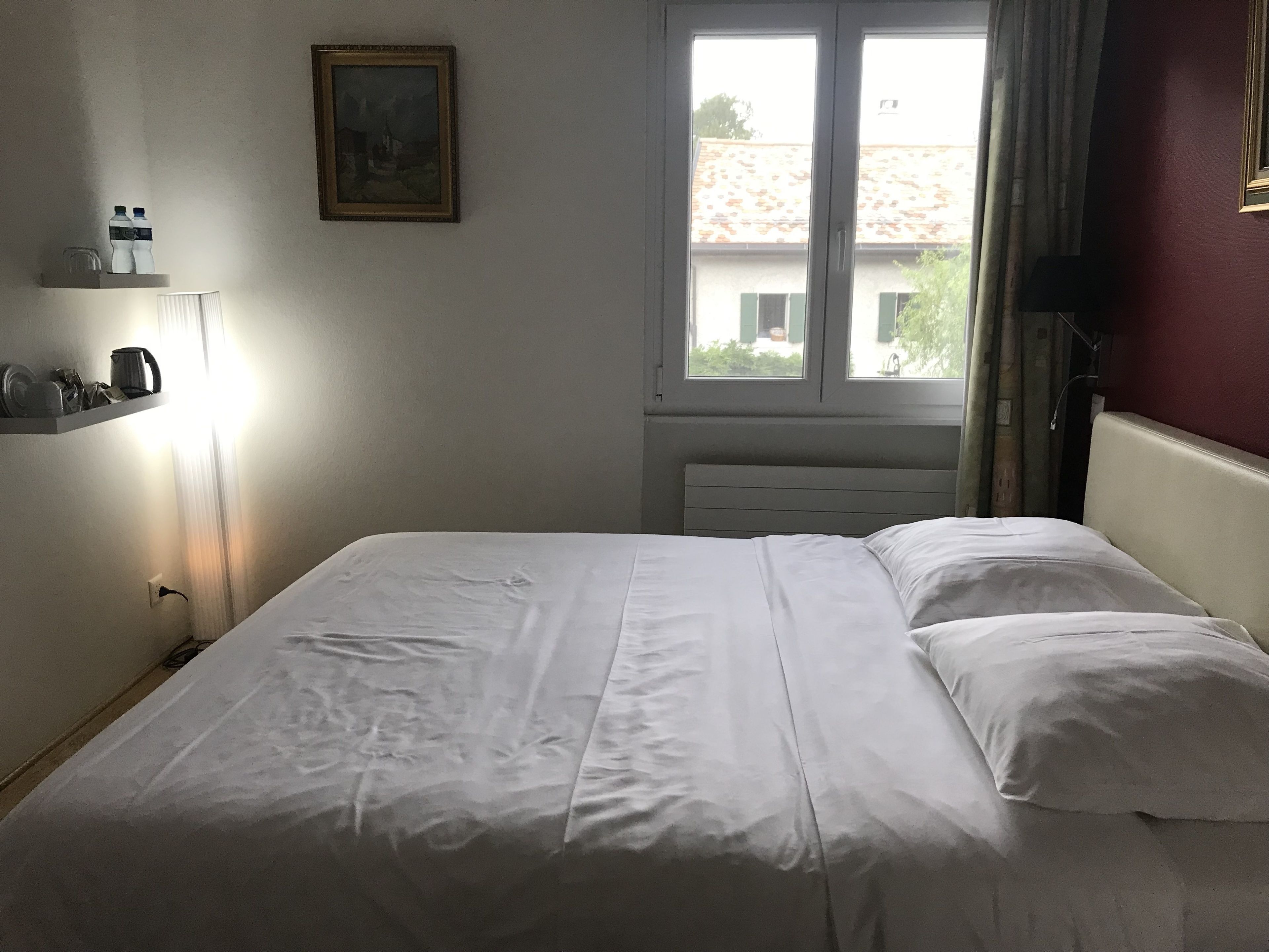 A neatly made bed with white linens, a small shelf with kitchen items, a painting on the wall, and a window with a view of buildings outside.