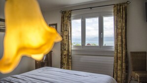 Un dormitorio con cama, ventana con cortinas, una silla y vista al mar.