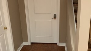 Un pasillo con una puerta blanca, piso de madera y una escalera al fondo.