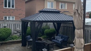 Un patio cubierto con mobiliario exterior, una pérgola y vista a un edificio residencial al fondo.